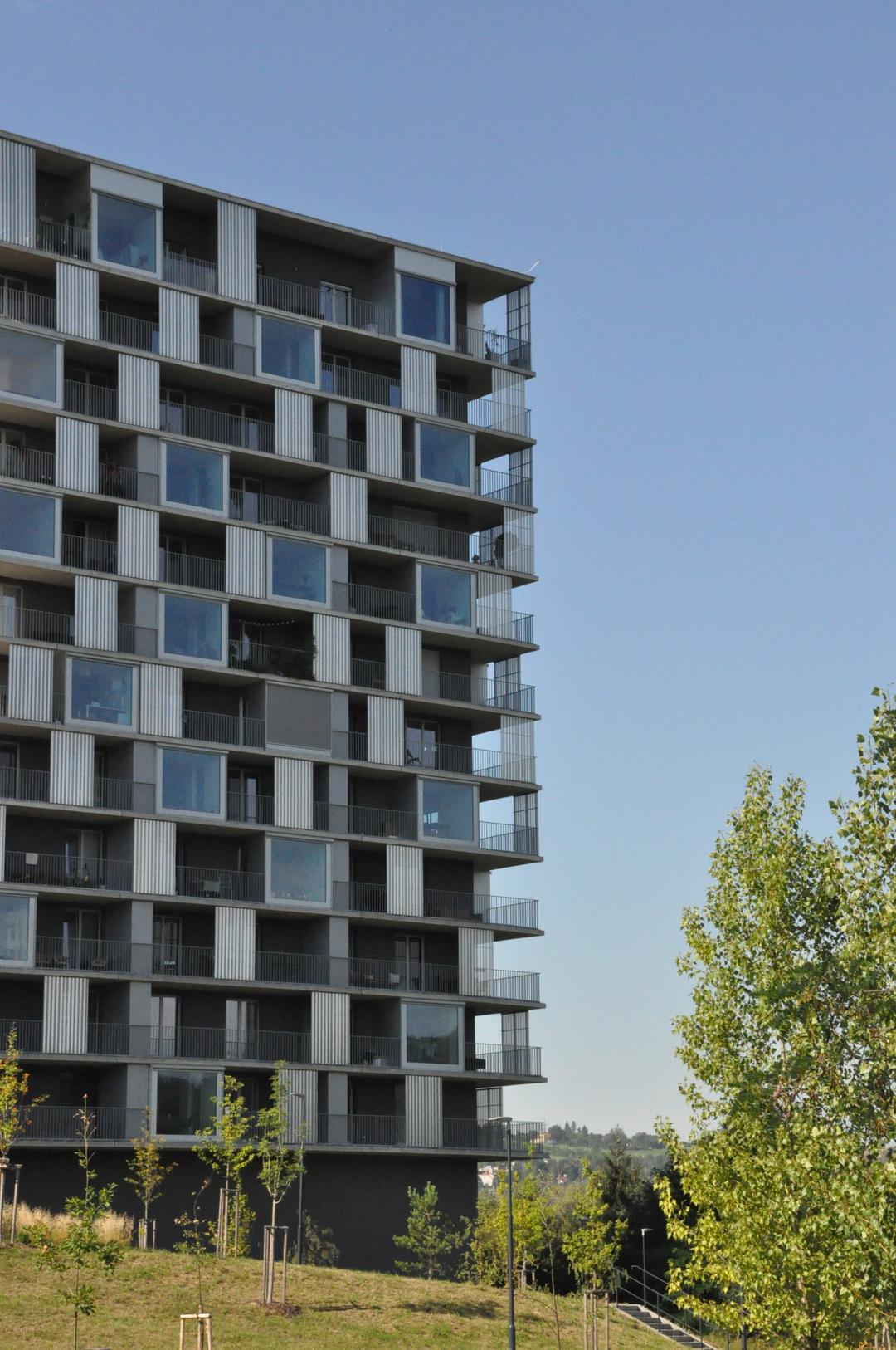 Ein Ausschnitt des modernen, mosaikartigen Gebäudekomplexes vor blauem Himmel, im Vordergrund Grünflächen und Bäume.