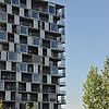 Ein Ausschnitt des modernen, mosaikartigen Gebäudekomplexes vor blauem Himmel, im Vordergrund Grünflächen und Bäume.