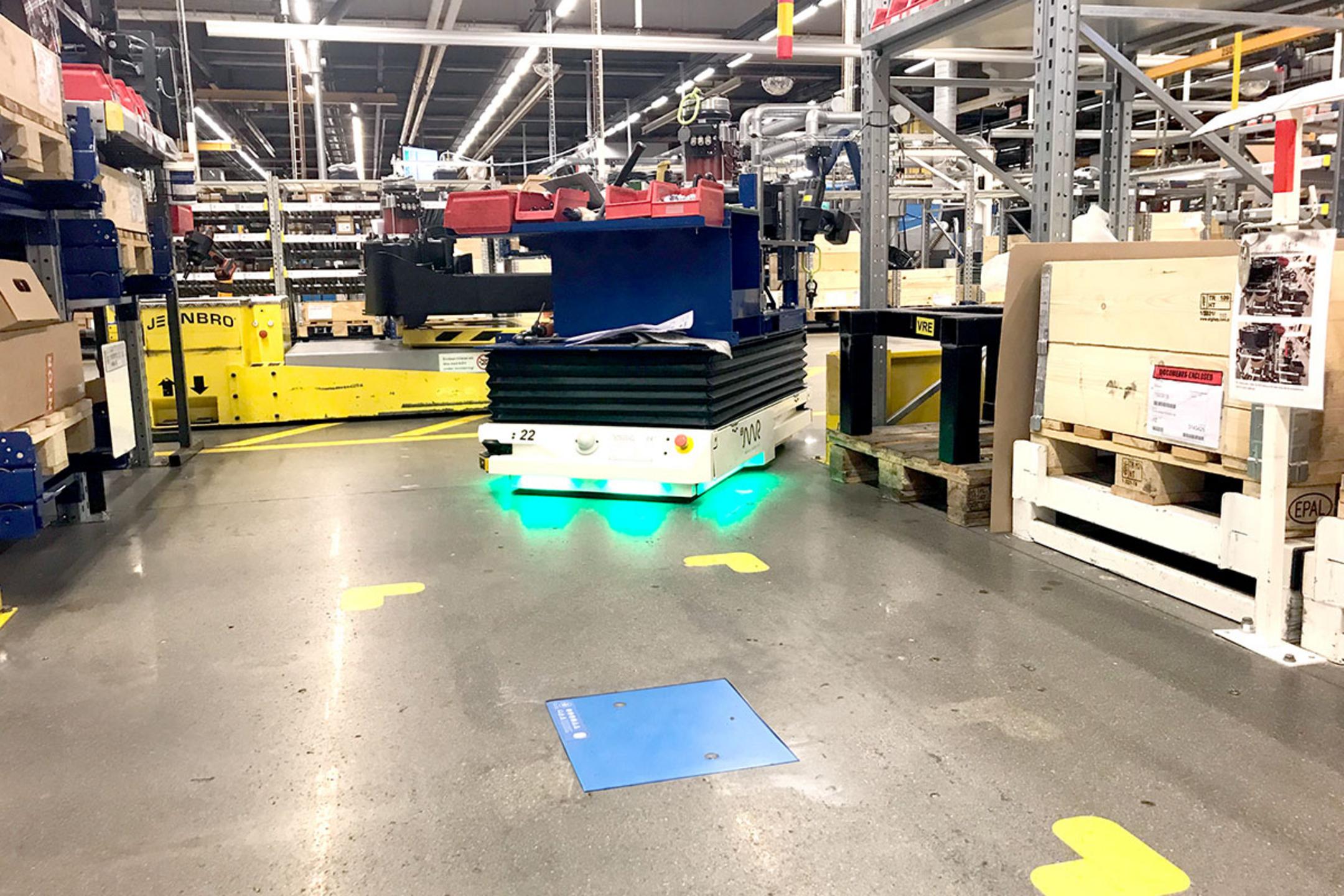 Automatisierter Transportroboter bewegt Waren durch ein Lager mit Regalen und Kartons, im Vordergrund befindet sich eine blaue induktive Ladeeinheit