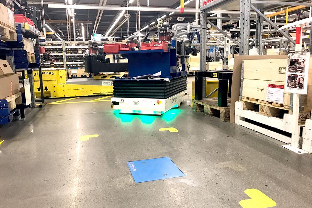 Automatisierter Transportroboter bewegt Waren durch ein Lager mit Regalen und Kartons, im Vordergrund befindet sich eine blaue induktive Ladeeinheit