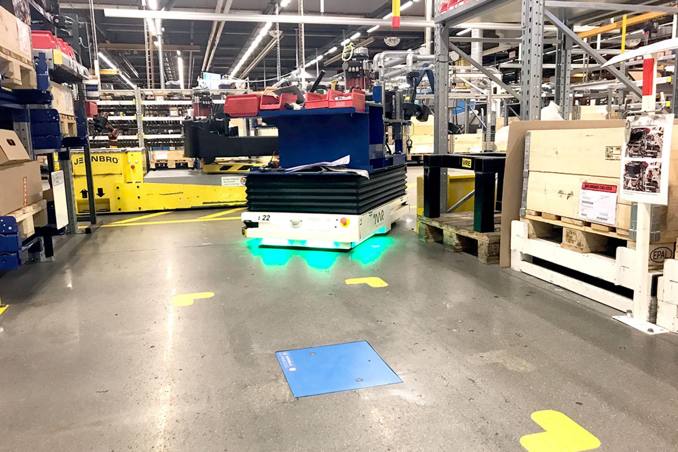 Automatisierter Transportroboter bewegt Waren durch ein Lager mit Regalen und Kartons, im Vordergrund befindet sich eine blaue induktive Ladeeinheit