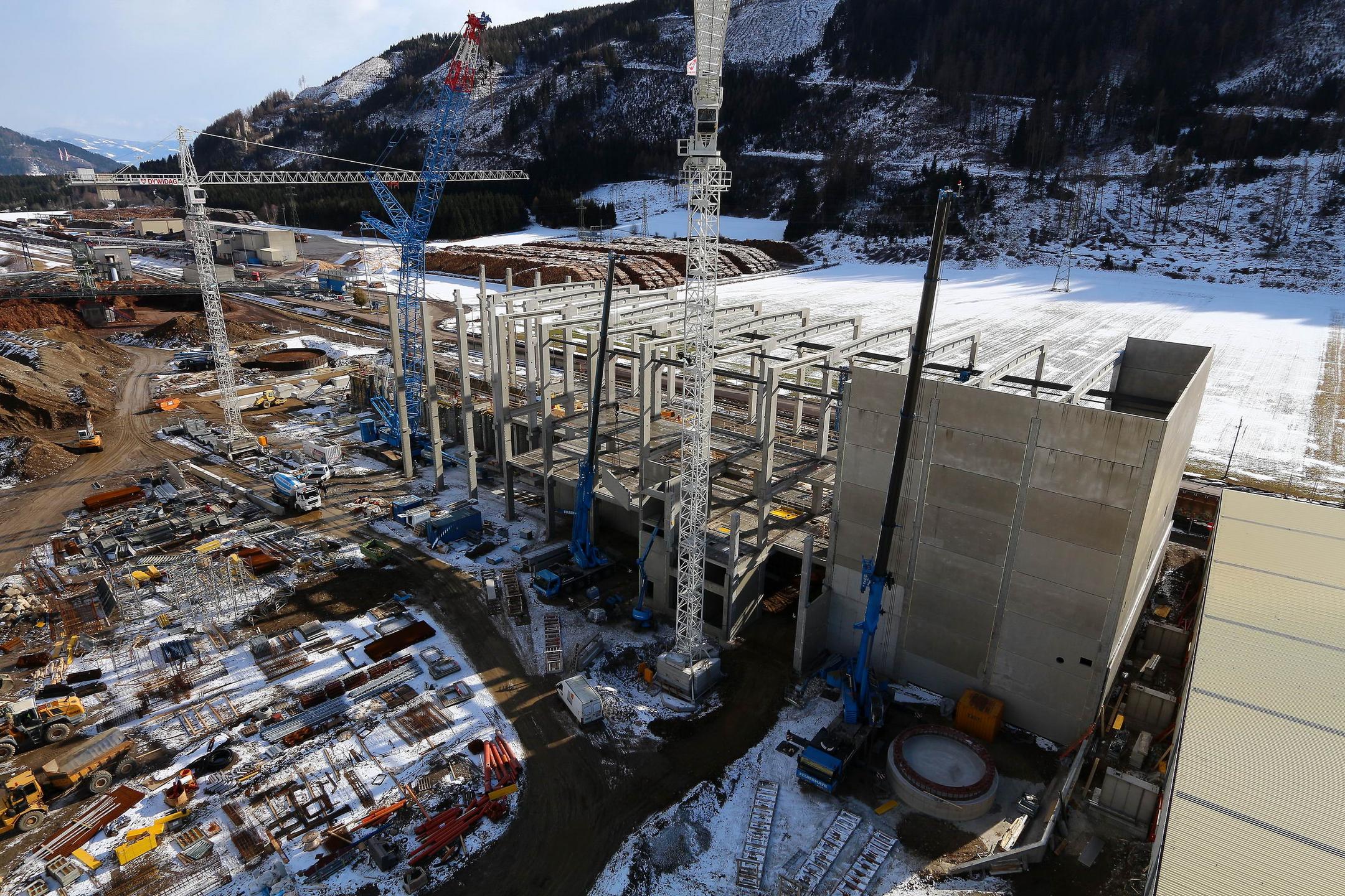 Große Baustelle im Gebirge mit Schnee, Kränen, Stahlgerüst, Betonwänden und Baumaschinen