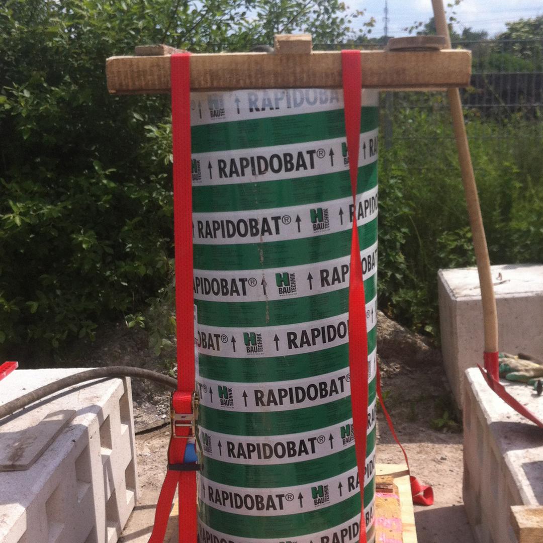 Nahaufnahme eines grün-weiß gestreiften Schalrohres mit Aufschrift "Rapidobat", befestigt mit roten Spanngurten, Baustellenumgebung