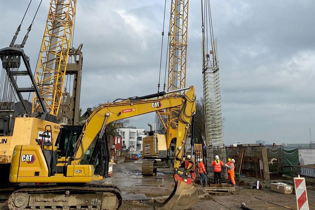 Baustelle mit Kran, Bagger und Bauarbeitern beim Einheben eines Bewehrungskorbs am Ufer unter bewölktem Himmel