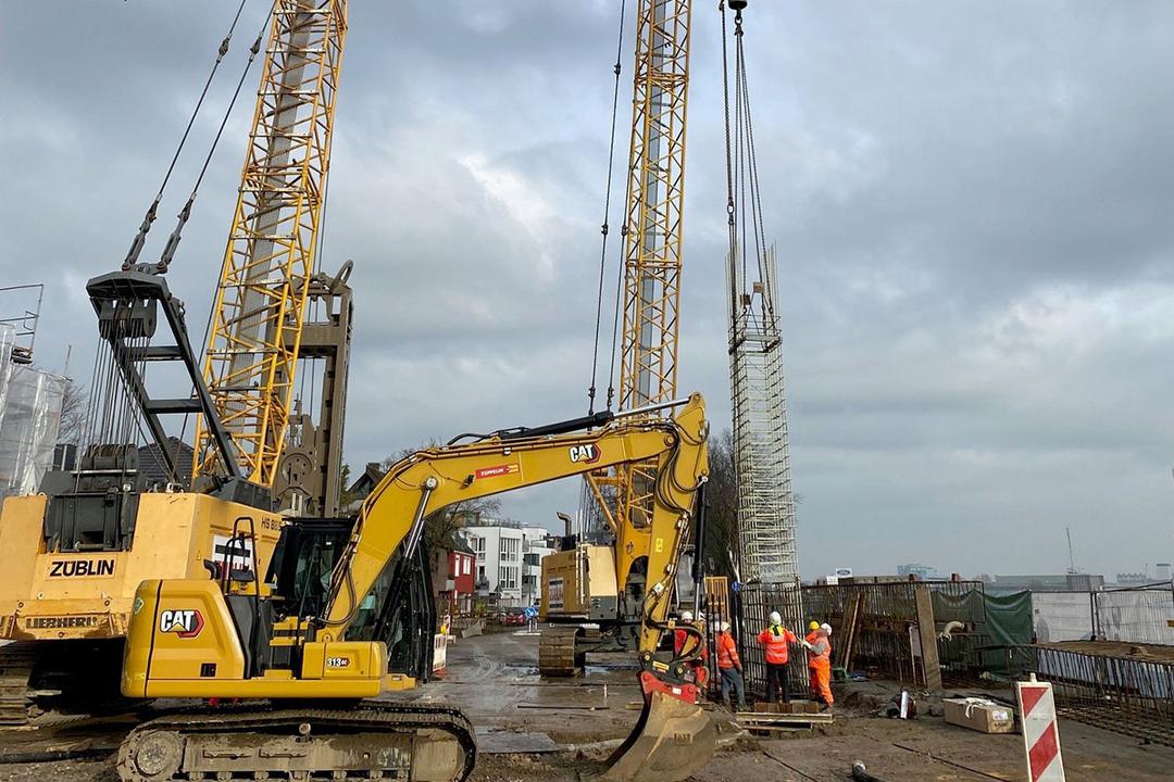 Baustelle mit Kran, Bagger und Bauarbeitern beim Einheben eines Bewehrungskorbs am Ufer unter bewölktem Himmel