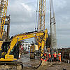 Baustelle mit Kran, Bagger und Bauarbeitern beim Einheben eines Bewehrungskorbs am Ufer unter bewölktem Himmel