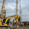 Baustelle mit Kran, Bagger und Bauarbeitern beim Einheben eines Bewehrungskorbs am Ufer unter bewölktem Himmel