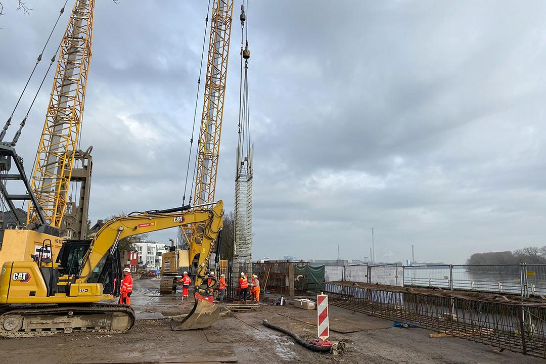 Bauarbeiter und Maschinen auf einer Baustelle am Wasser, ein Kran hebt einen Bewehrungskorb in die Höhe