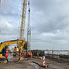 Bauarbeiter und Maschinen auf einer Baustelle am Wasser, ein Kran hebt einen Bewehrungskorb in die Höhe