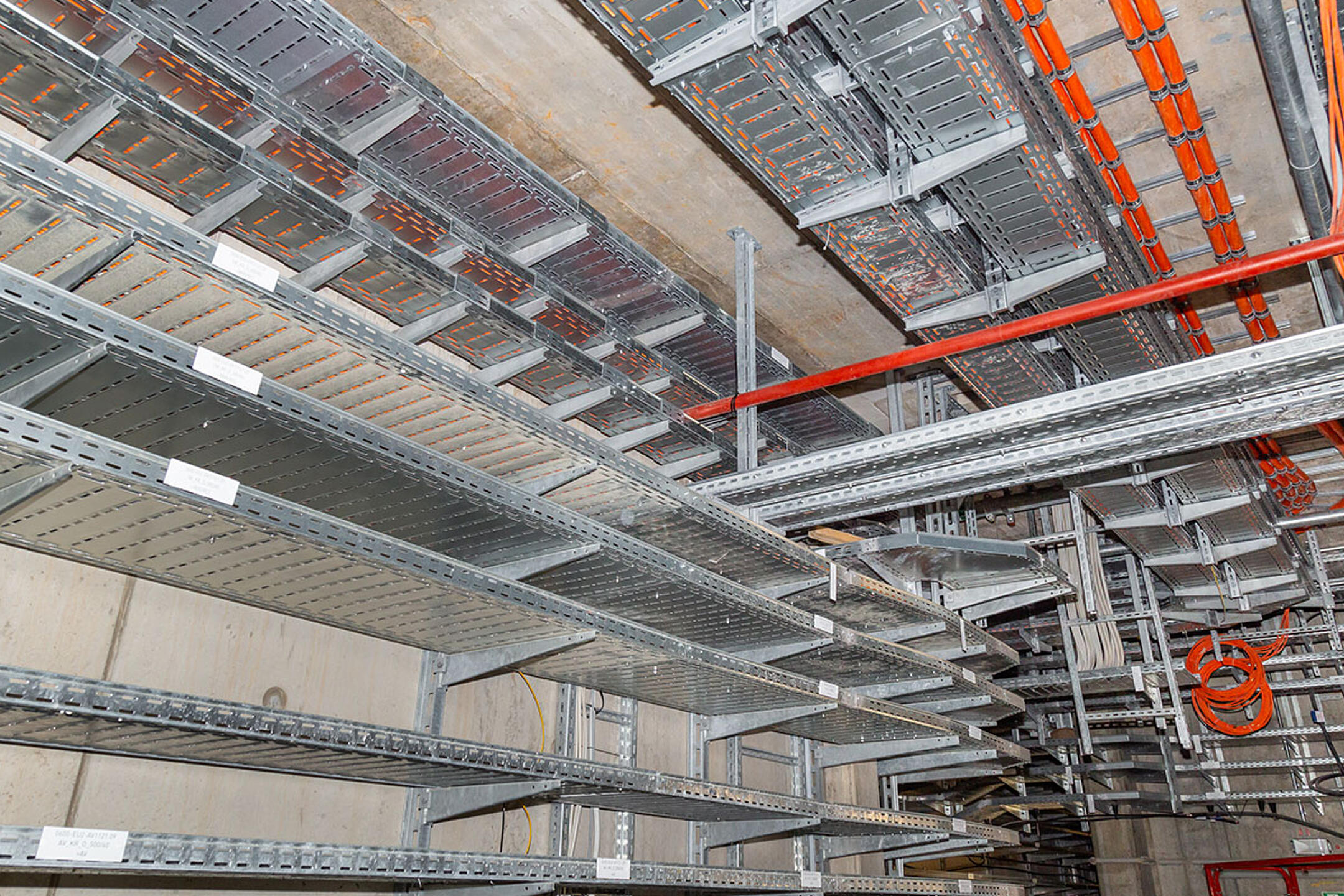 Kabelrinnen mit orangefarbenen Kabeln befestigt an Betondecke und Betonwand in einem Technikraum