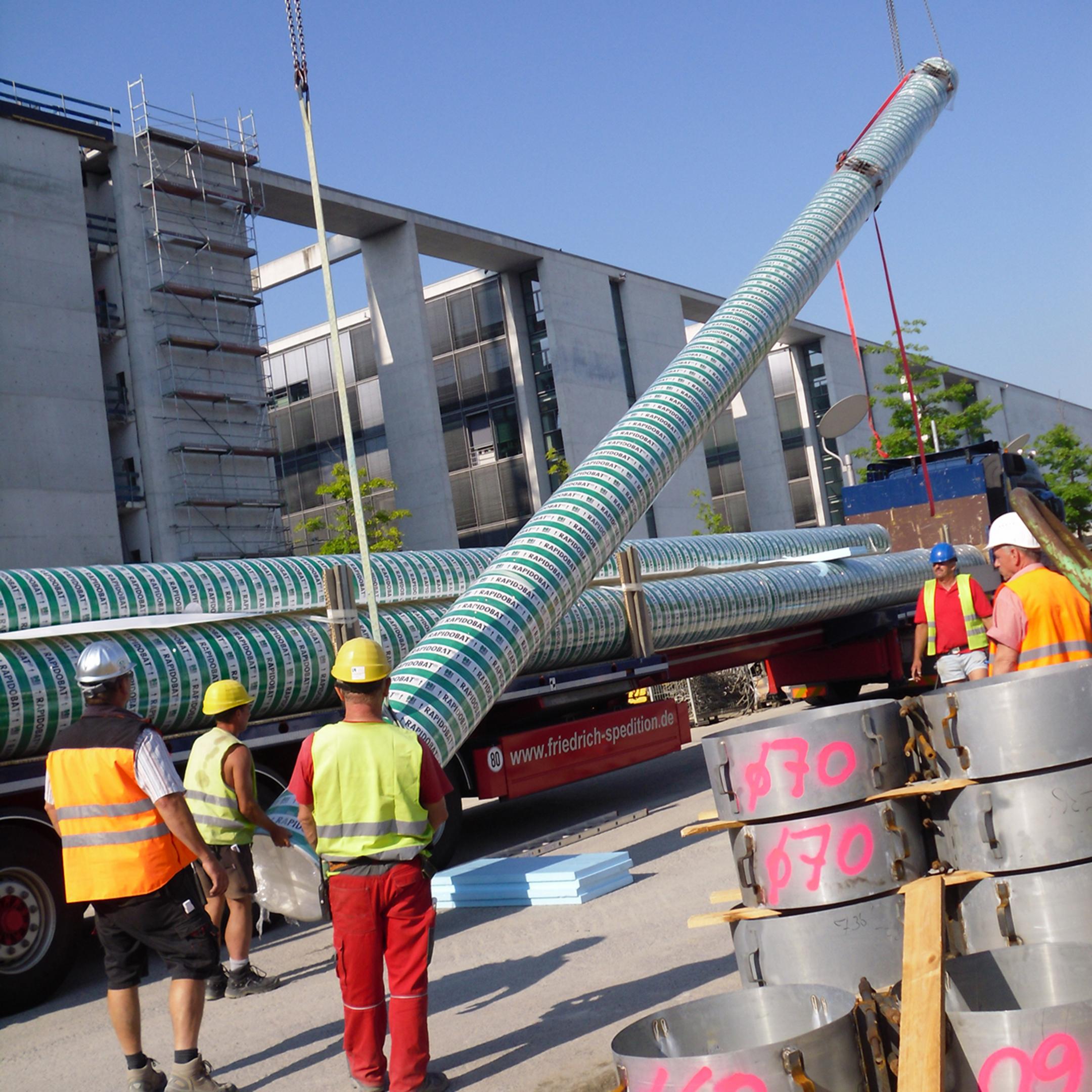 Bauarbeiter mit Westen und Helmen entladen mithilfe eines Krans einen LKW, der Schalrohre liefert. Im Hintergrund ist die ein Gebäude zu sehen.