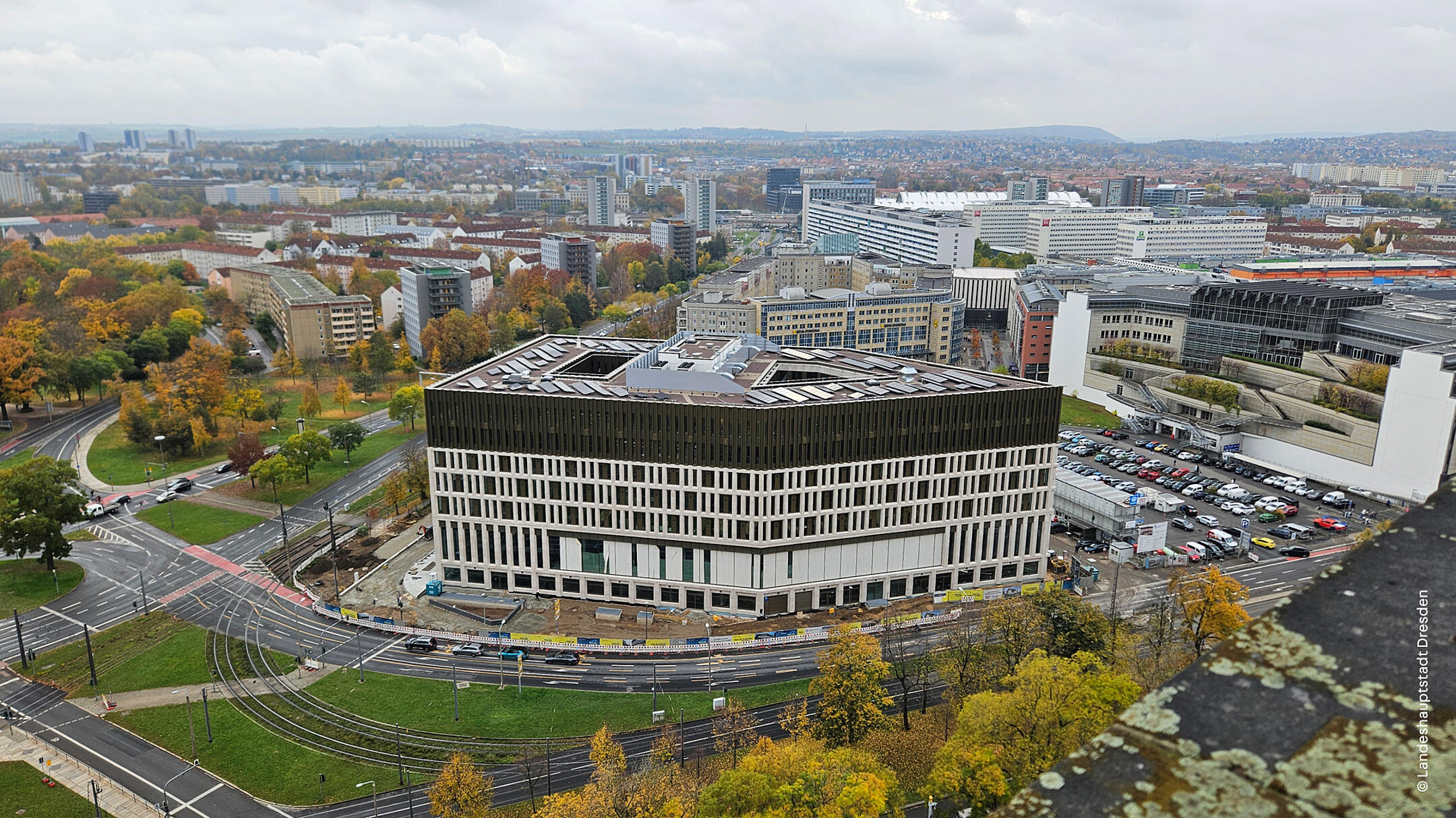 Luftaufnahme eines Gebäudes, das von einer Baustelle umgeben ist, ein Parkplatz angrenzend. Im Hintergrund sieht man weitere Gebäude der Stadt und Bäume.