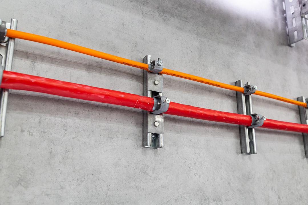Orange und rote Leitungen an einer Betonwand mit Kabelschellen und Montageschienen zur geordneten Leitungsführung.