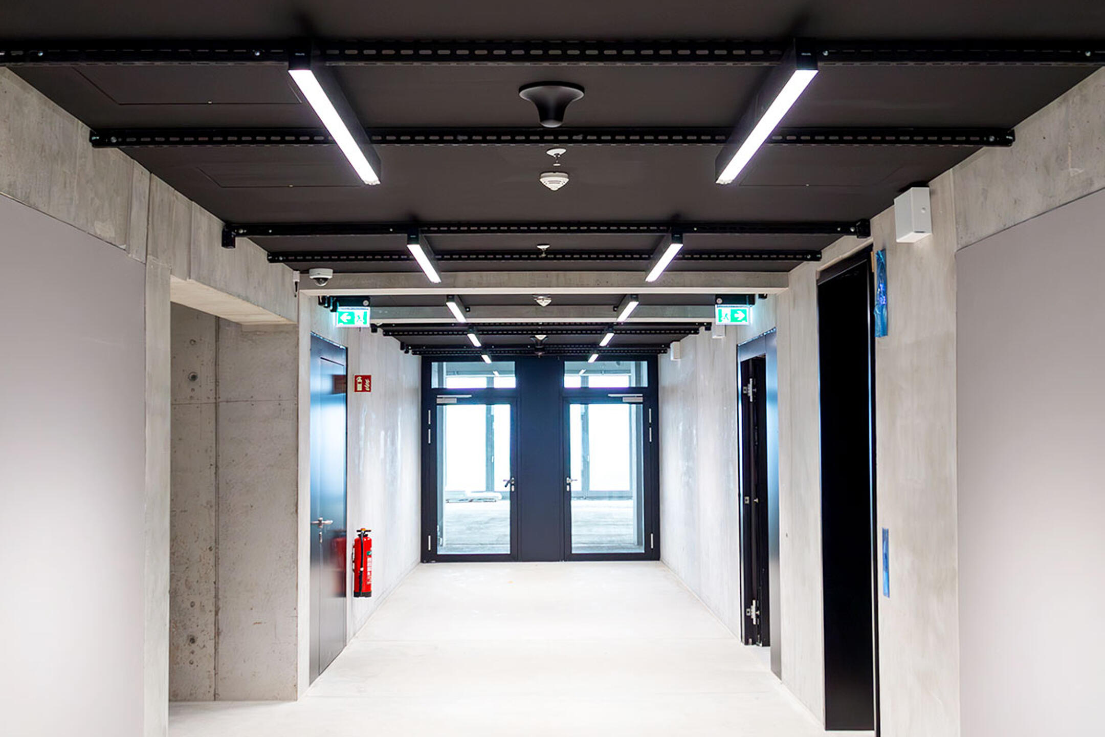 Heller Flur mit Betonwänden, schwarzen Türen, einer schwarzen Decke mit weißen LED-Lichtern und Ausgangstüren am Ende