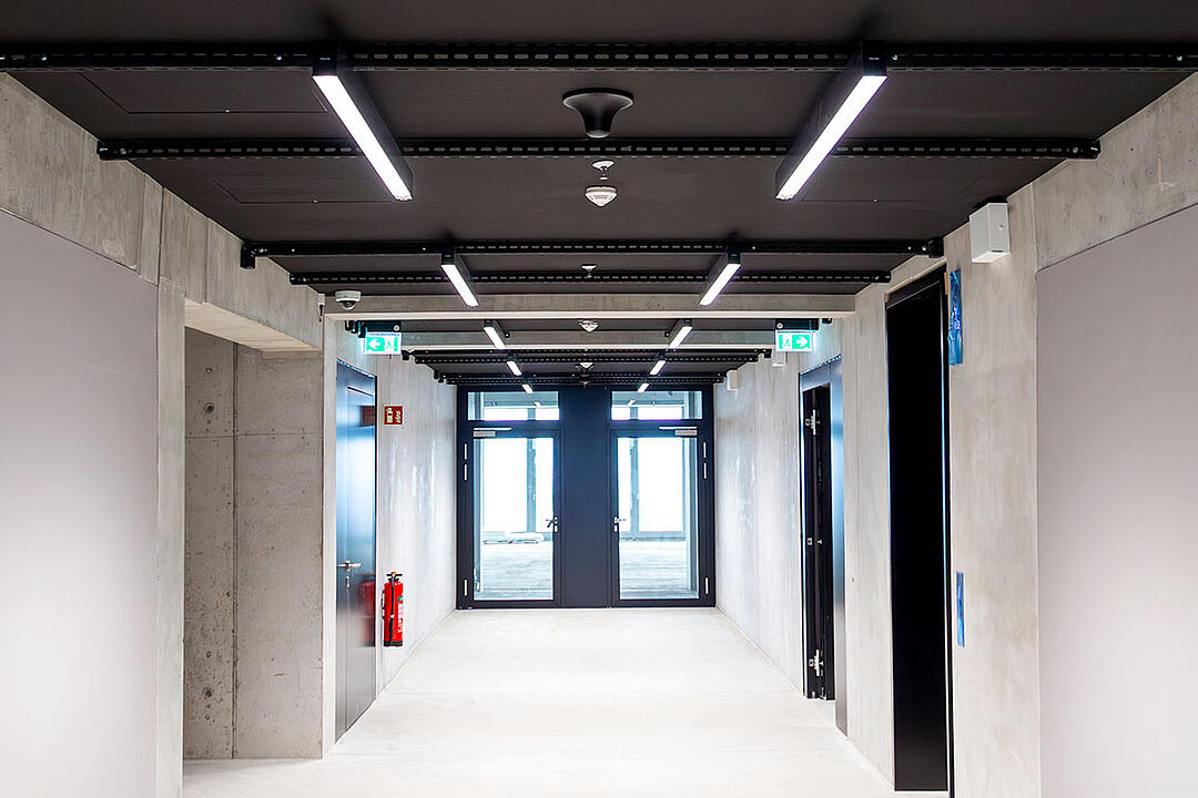Heller Flur mit Betonwänden, schwarzen Türen, einer schwarzen Decke mit weißen LED-Lichtern und Ausgangstüren am Ende