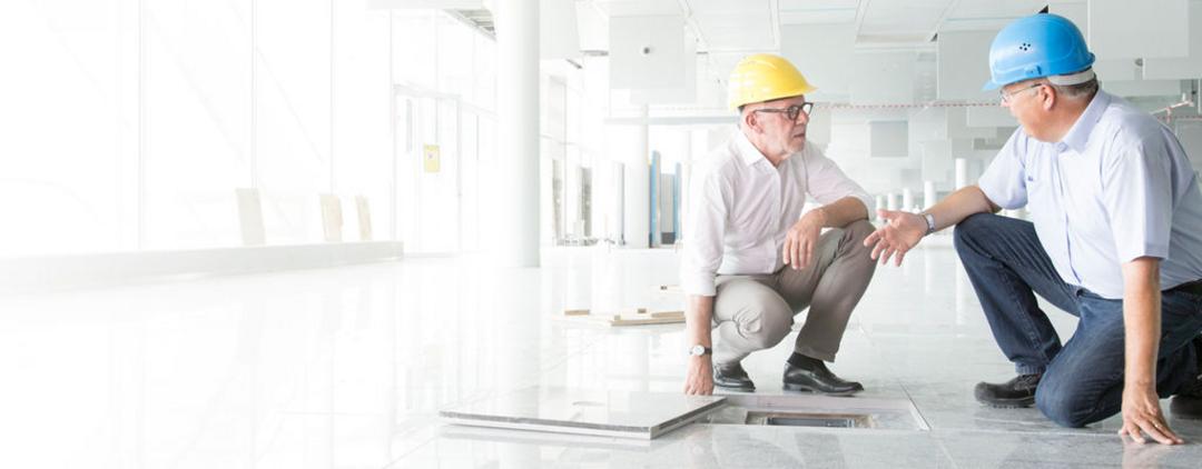 Zwei Personen mit Bauhelmen hocken in einem Gebäude am Boden und unterhalten sich freundlich.