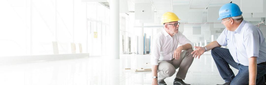 Zwei Personen mit Bauhelmen hocken in einem Gebäude am Boden und unterhalten sich freundlich.