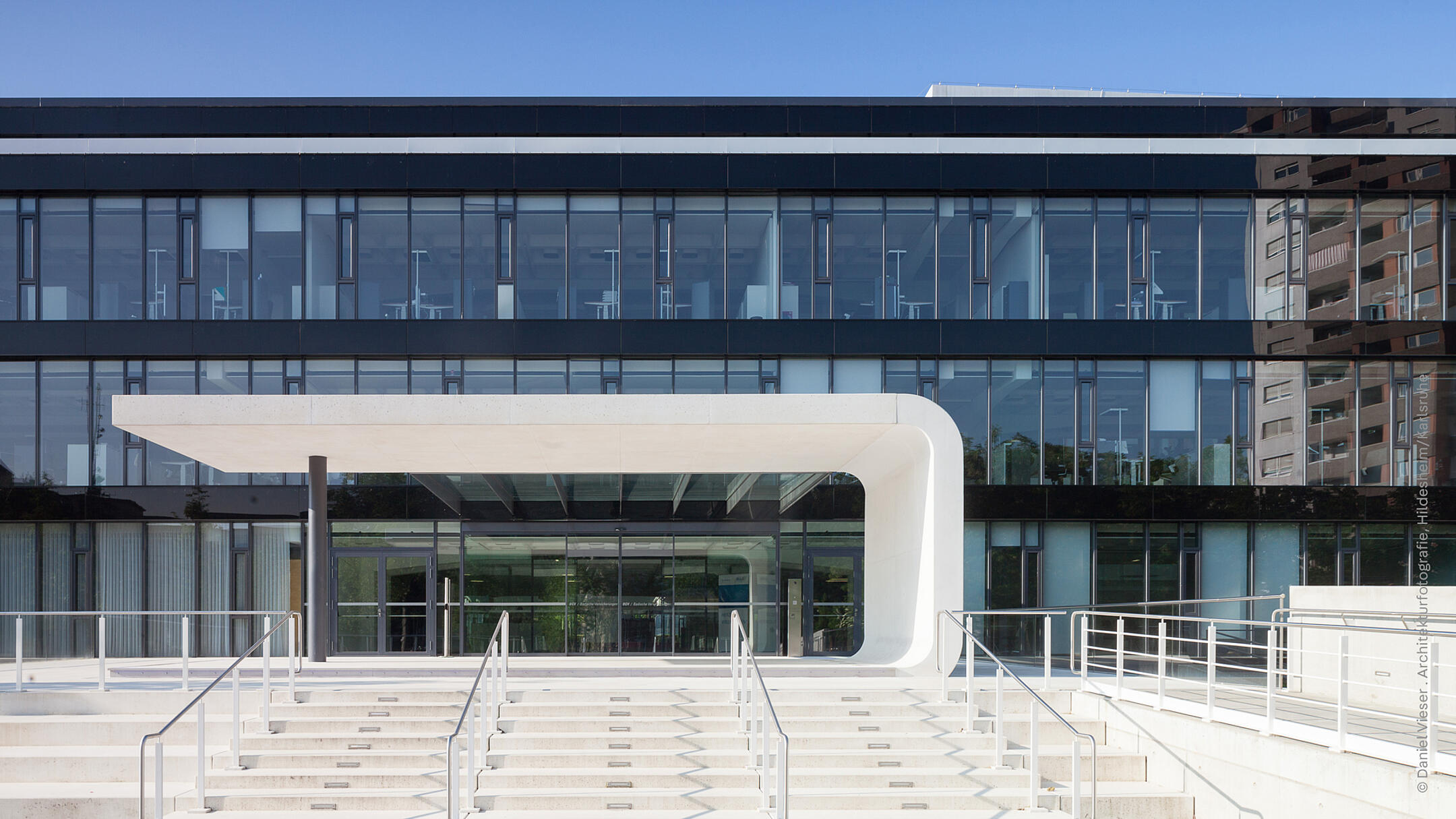 Zentraler Bildinhalt ist ein schwarzes Gebäude mit einer Glasfront, im Hintergrund ist ein blauer Himmel zu erkennen. Weiße Stufen und eine weiße massive Überdachung prägen den Eingangsbereich.