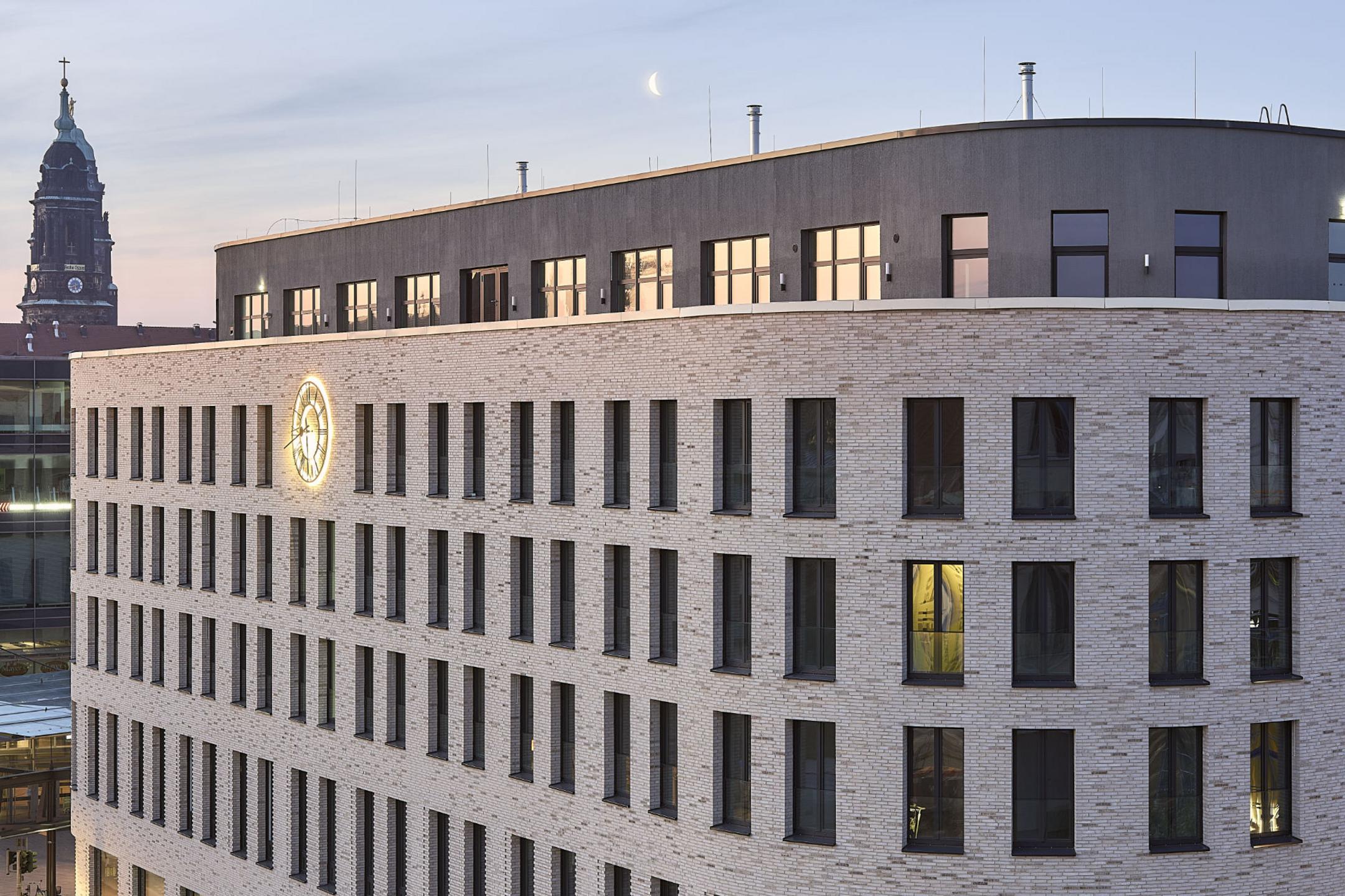 Aufnahme der oberen Stockwerke des Haus Postplatz in Dresden mit einer beleuchteten Uhr auf der Fassade und einem Kurchturm im Hintergrund