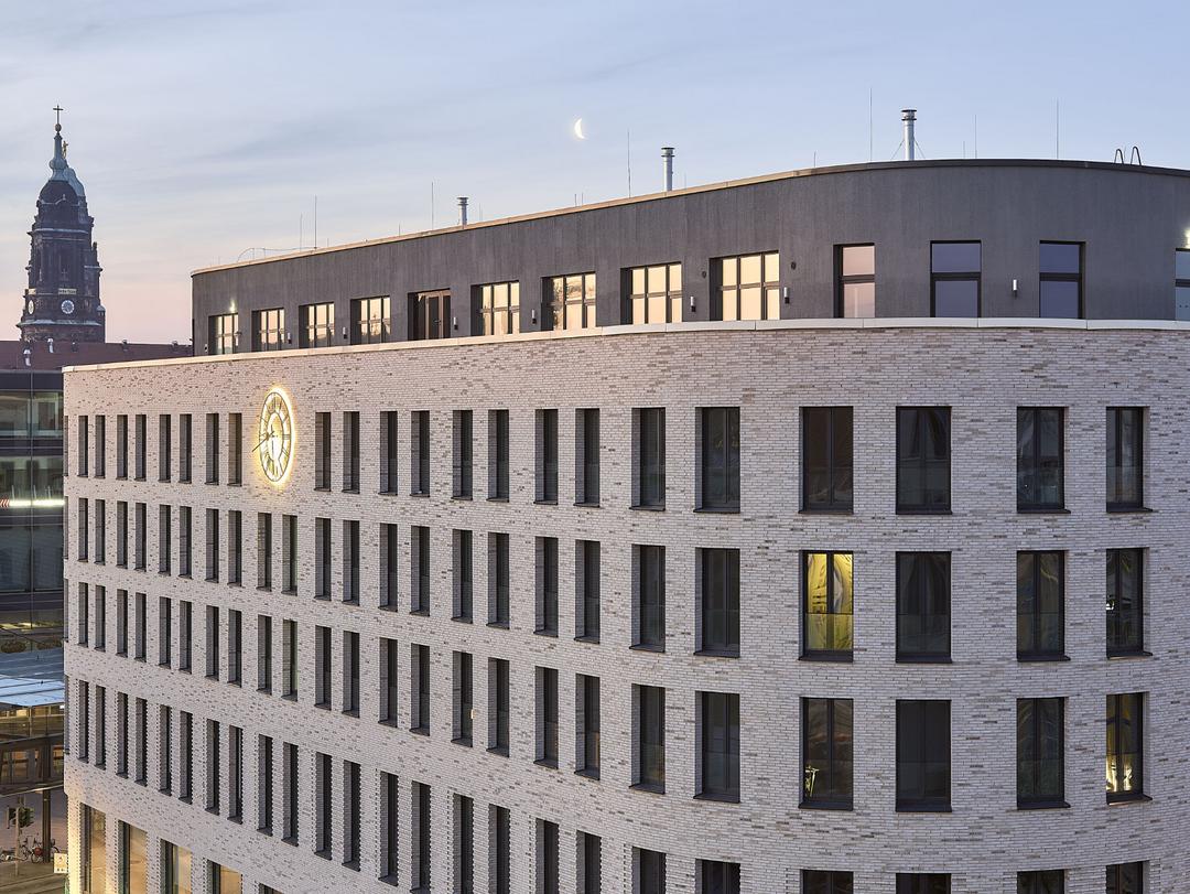 Aufnahme der oberen Stockwerke des Haus Postplatz in Dresden mit einer beleuchteten Uhr auf der Fassade und einem Kurchturm im Hintergrund