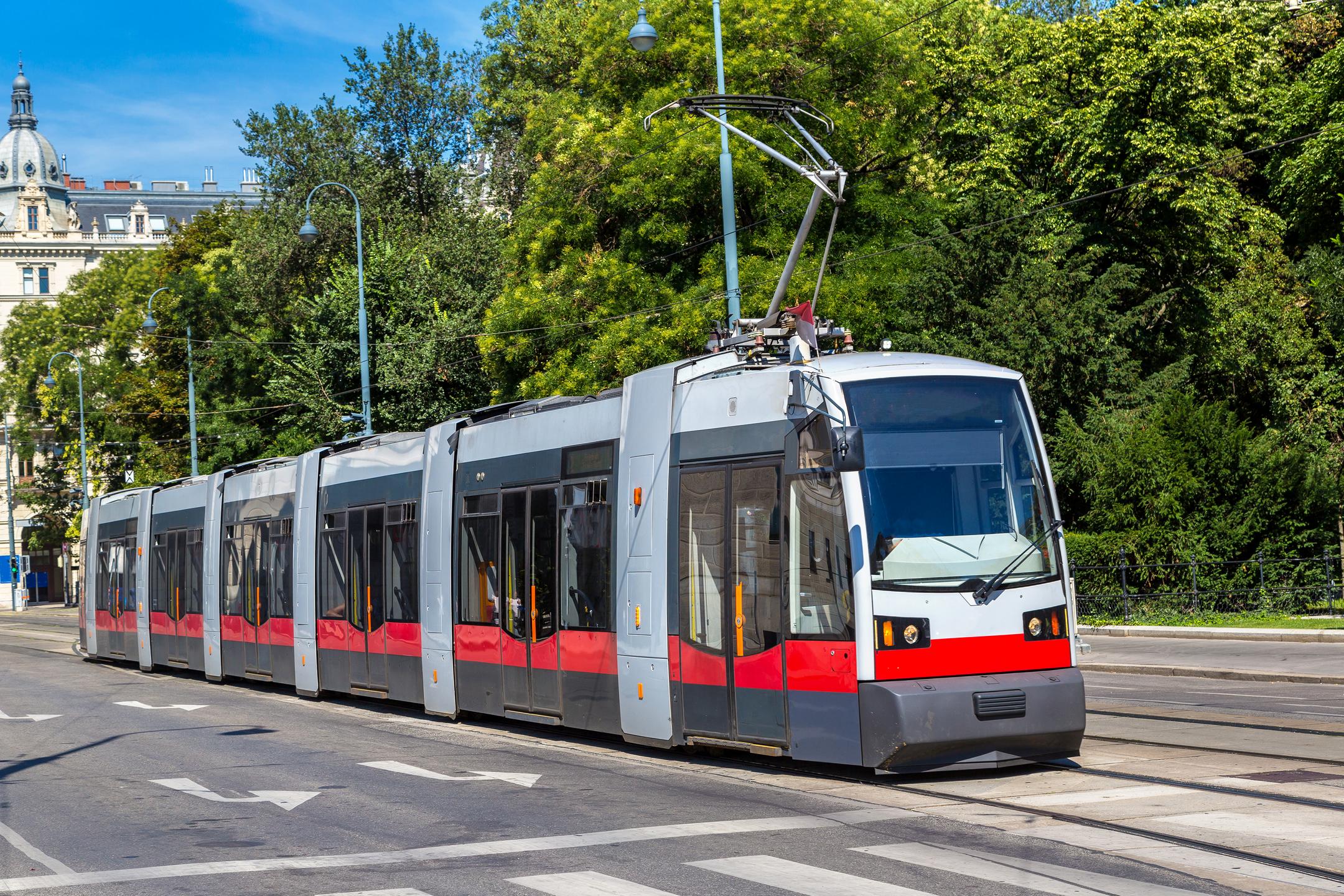 Grau-rote Straßenbahn vor einem begrünten Hintergrund, im Anschnitt sind eine Gebäude und blauer Himmel zu erkennen.