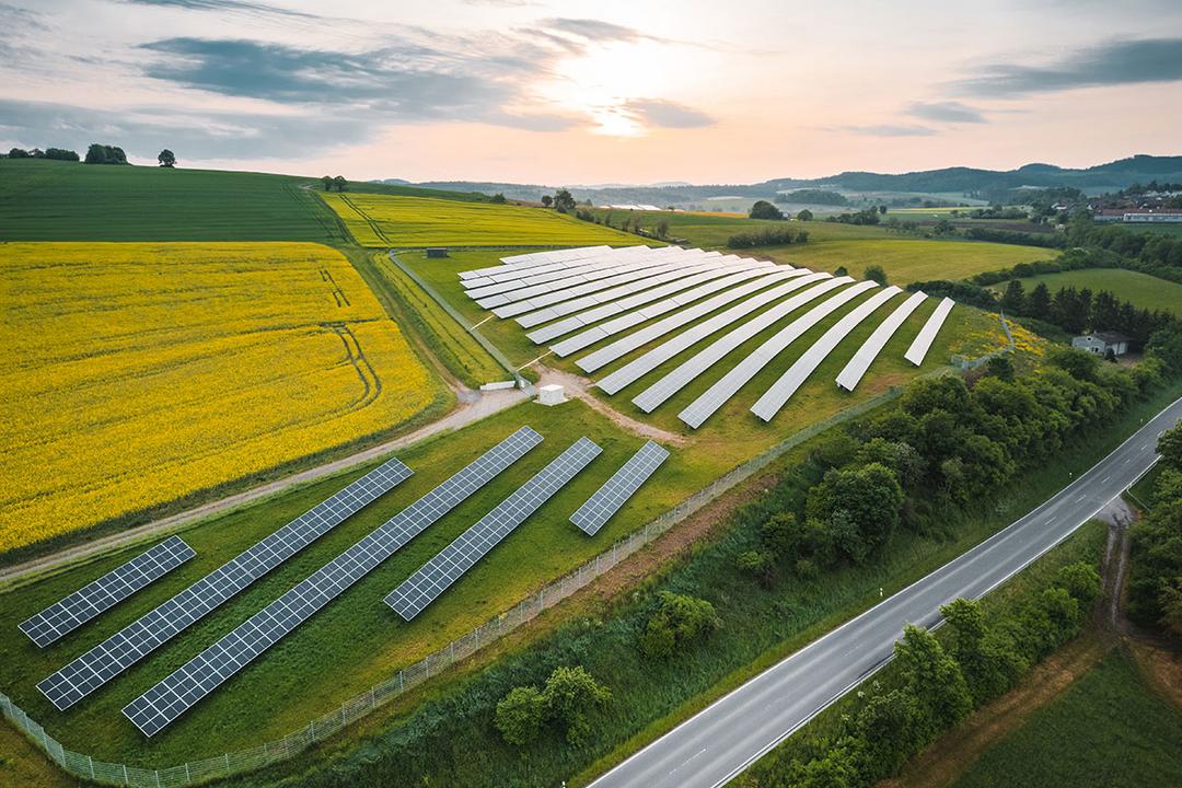Luftaufnahme einer großen Solaranlage auf Hügeln mit Feldern, Straße daneben, Sonnenaufgang und teils bewölktem Himmel