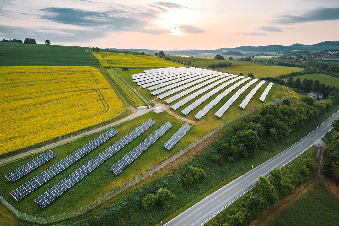 Luftaufnahme einer großen Solaranlage auf Hügeln mit Feldern, Straße daneben, Sonnenaufgang und teils bewölktem Himmel