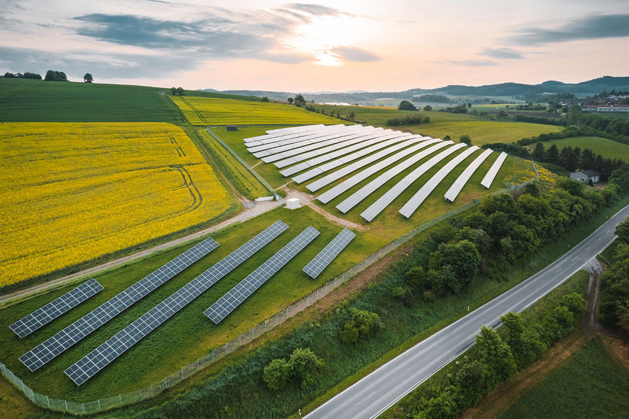 Luftaufnahme einer großen Solaranlage auf Hügeln mit Feldern, Straße daneben, Sonnenaufgang und teils bewölktem Himmel