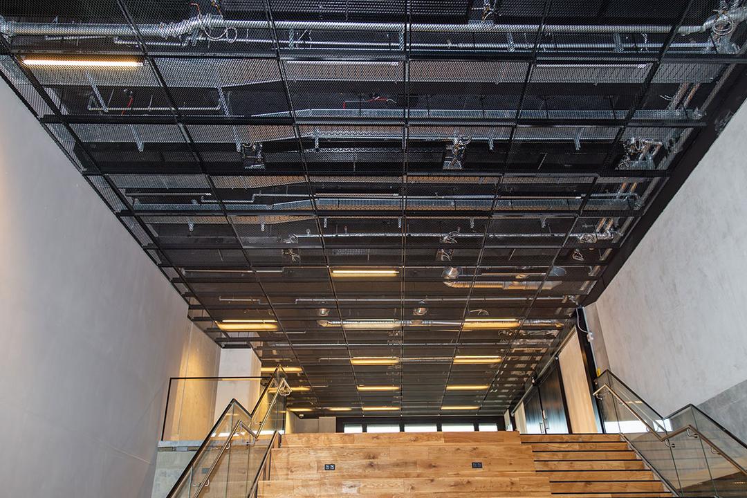 Innenaufnahme eines Treppenhauses mit Holzstufen und einer schwarzen Gitterdecke mit Kabelrinnen, sichtbar sind Leitungen und Beleuchtung