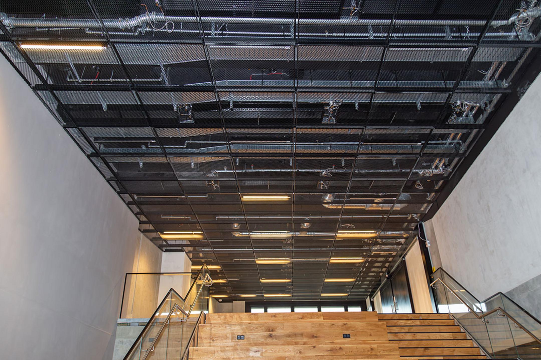 Innenaufnahme eines Treppenhauses mit Holzstufen und einer schwarzen Gitterdecke mit Kabelrinnen, sichtbar sind Leitungen und Beleuchtung