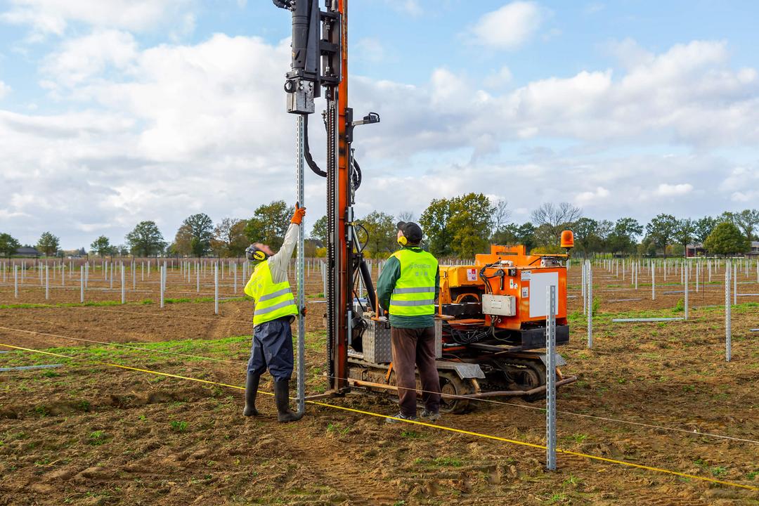 Zwei Arbeiter installieren mit einer Rammmaschine Stahlpfosten für eine PV-Unterkonstruktion auf einem freien Feld.