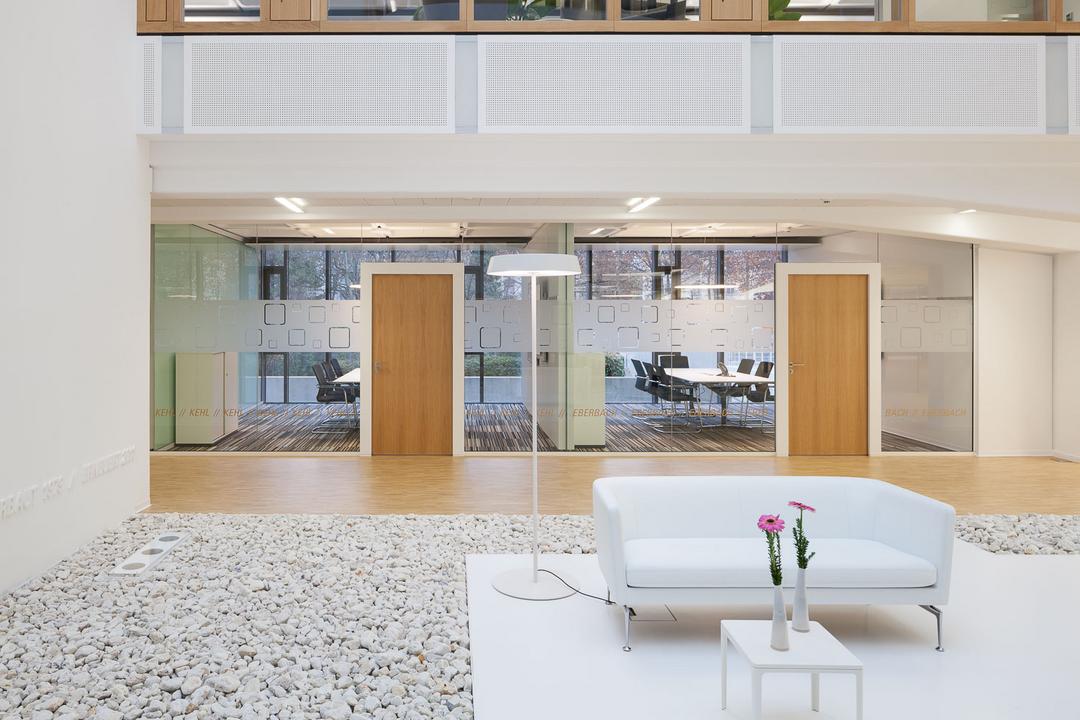 Ein moderner, einladender Eingangs- und Aufenthaltsbereich in einem Bürogebäude, mit Sitzgelegenheiten und verglasten Büros im Hintergrund.