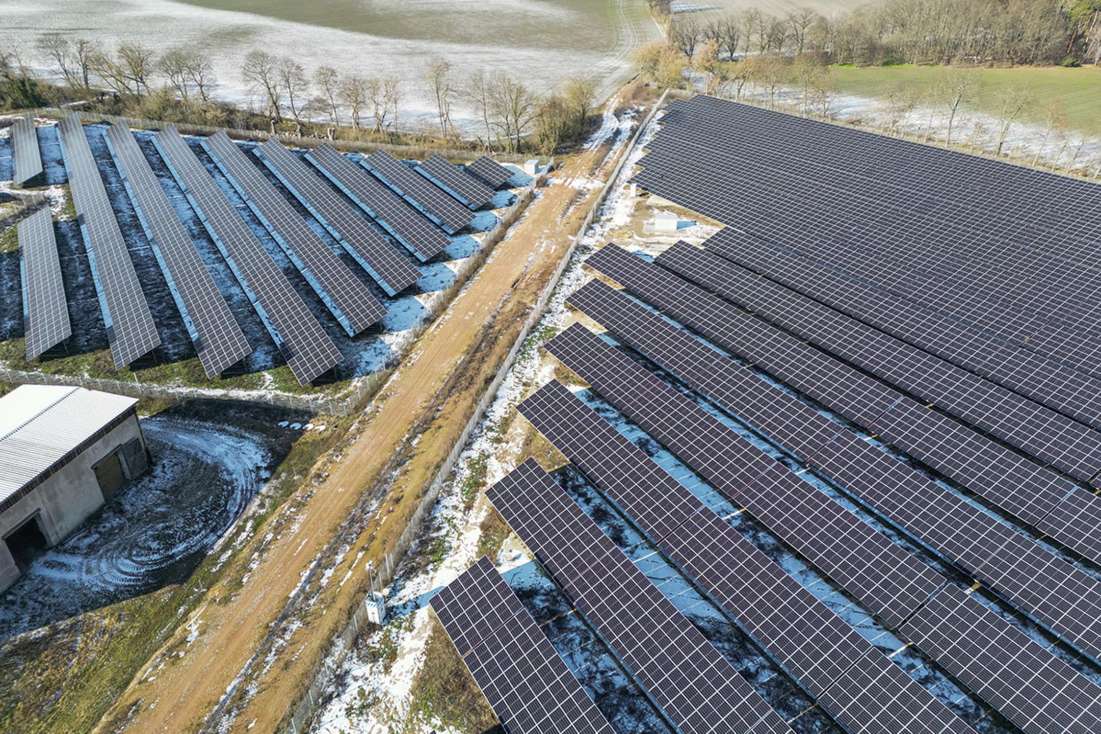 Luftaufnahme eines Solarparks, durch den ein Feldweg führt, Felder grenzen an und sind teilweise von Schnee bedeckt