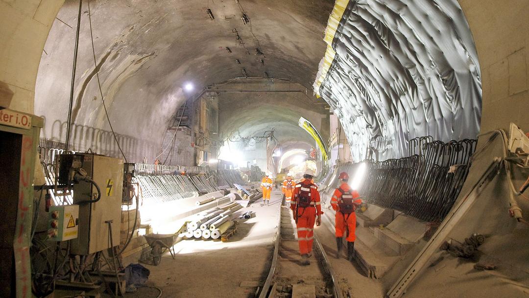 Tunnelbaustelle mit Bauarbeitern in Schutzkleidung, Schienen, Bewehrung und Baustoffen im unterirdischen Rohbau.