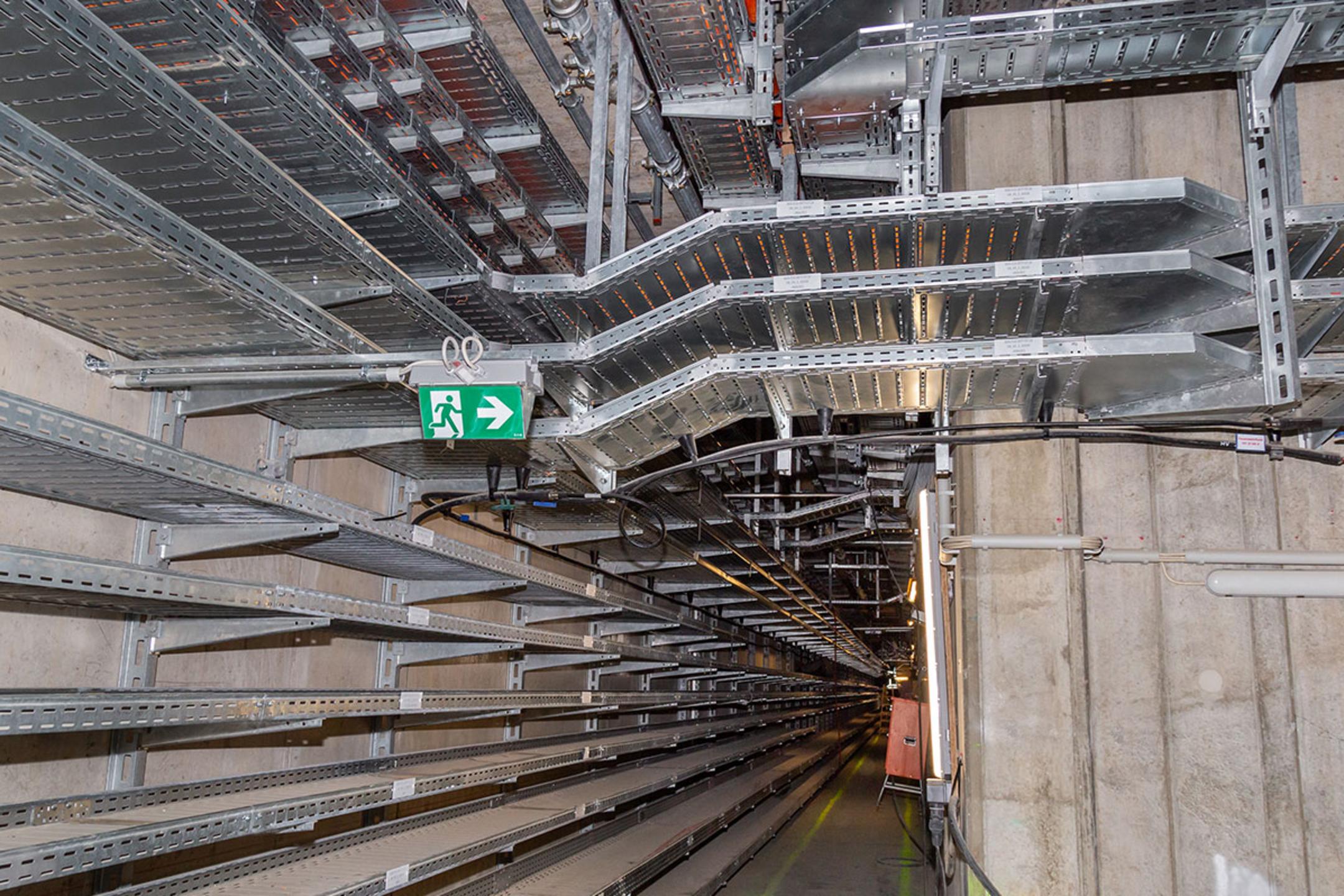 Langer Gang mit Kabelrinnen an der Wand und an der Decke, Notausgangszeichen und Betonwänden in einem Technik- oder Versorgungstunnel