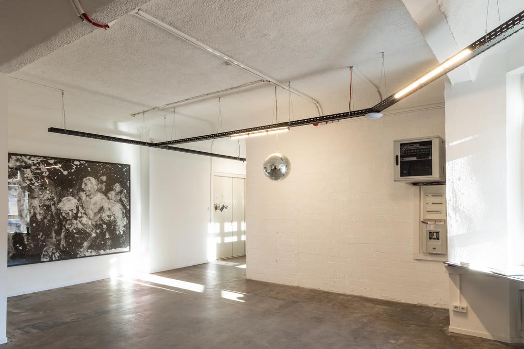 Totalansicht eines Raumes mit einem Bild in der linken Bildhälfte. Von der Decke hängen schwarze Kabelrinnen, an denen Leuchten und eine Discokugel befestigt sind.