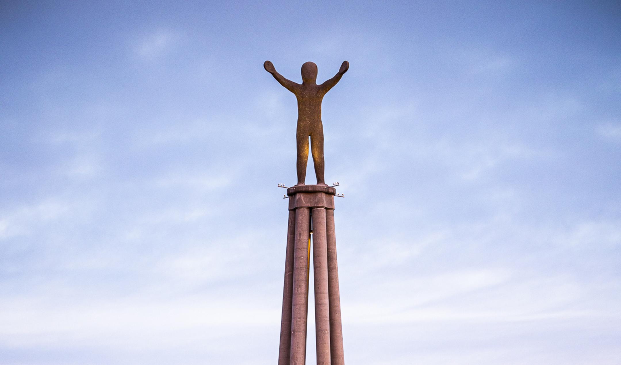Monument mit erhobener Statue auf Säulenplattform vor blauem Himmel mit vereinzelten Wolken