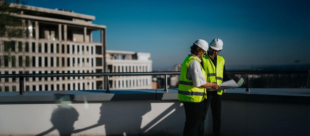 Zwei Personen mit gelben Warnwesten und Bauhelmen mit PohlCon-Logo stehen auf dem Dach eines Gebäudes und sehen sich einen Plan an.