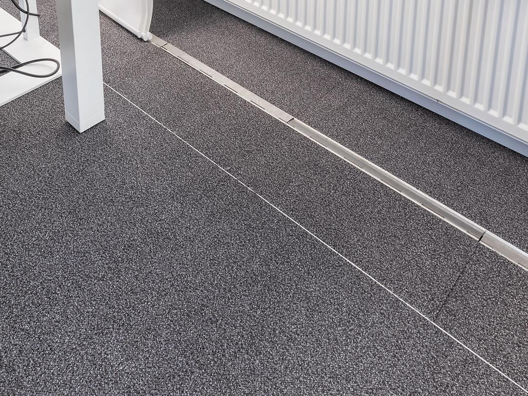 Ein teppichbedeckter Büroboden, durch den ein unauffälliges, estrichbündiges Kanalsystem für Kabel verläuft, im Hintergrund eine Heizung.