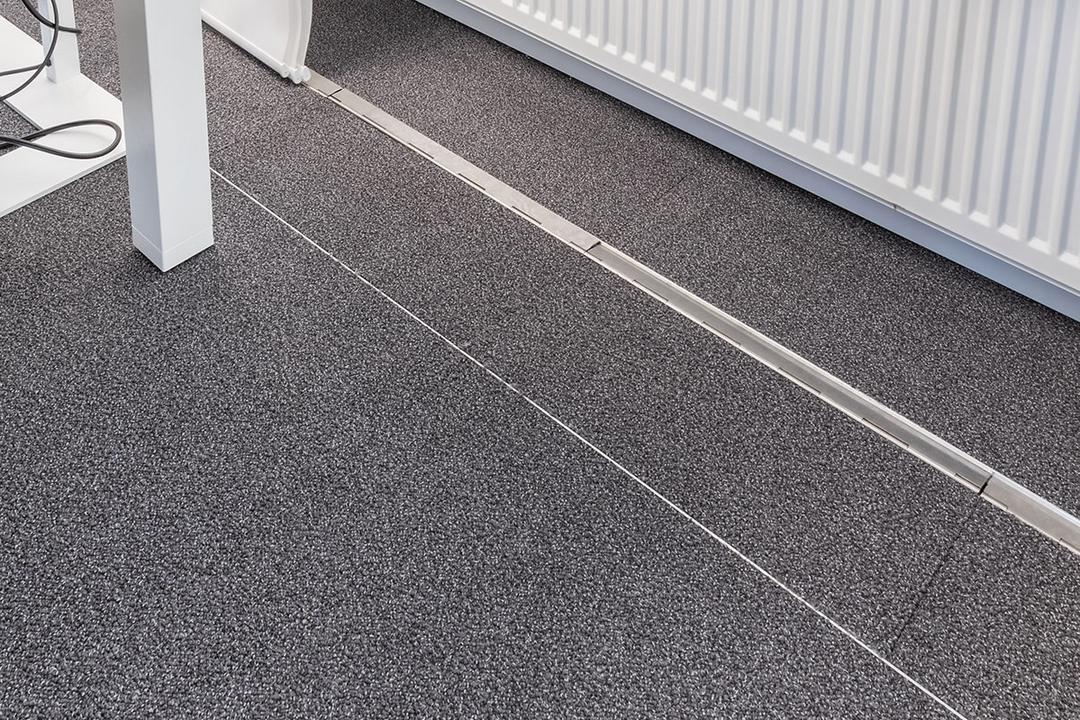 Ein teppichbedeckter Büroboden, durch den ein unauffälliges, estrichbündiges Kanalsystem für Kabel verläuft, im Hintergrund eine Heizung.
