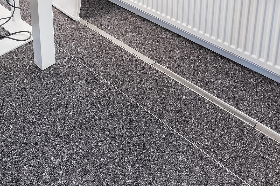 Ein teppichbedeckter Büroboden, durch den ein unauffälliges, estrichbündiges Kanalsystem für Kabel verläuft, im Hintergrund eine Heizung.