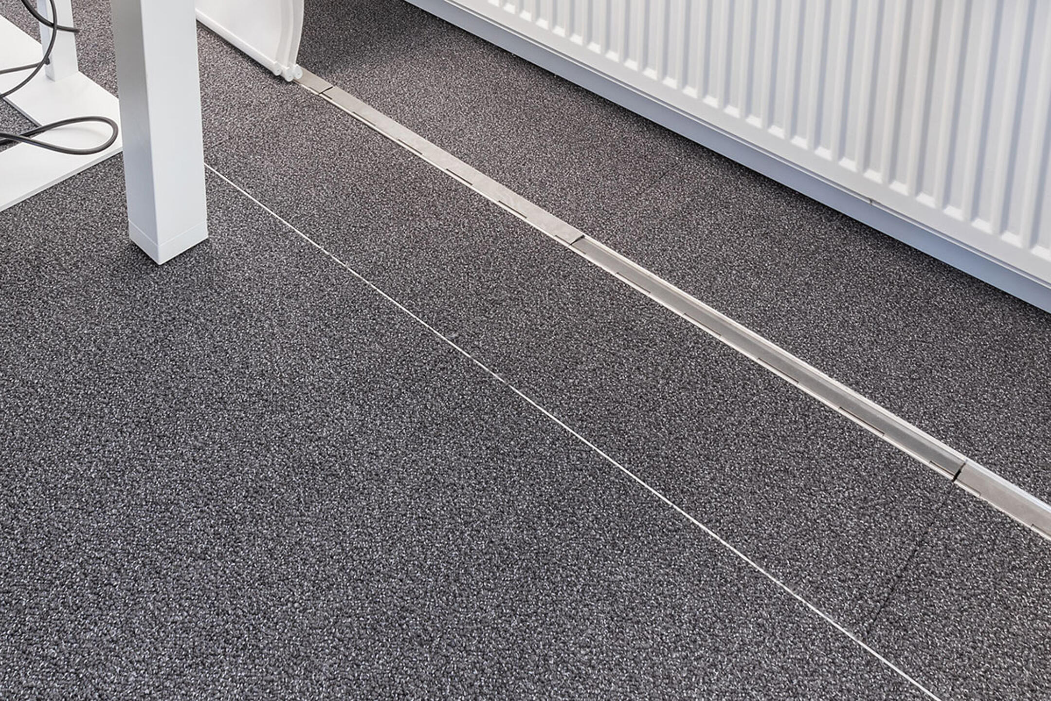 Ein teppichbedeckter Büroboden, durch den ein unauffälliges, estrichbündiges Kanalsystem für Kabel verläuft, im Hintergrund eine Heizung.