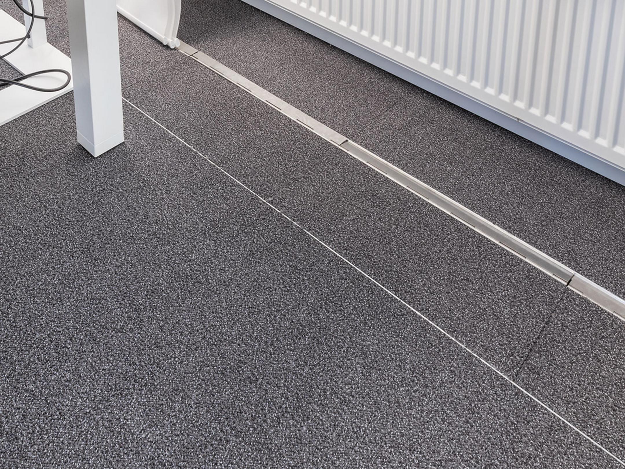Ein teppichbedeckter Büroboden, durch den ein unauffälliges, estrichbündiges Kanalsystem für Kabel verläuft, im Hintergrund eine Heizung.