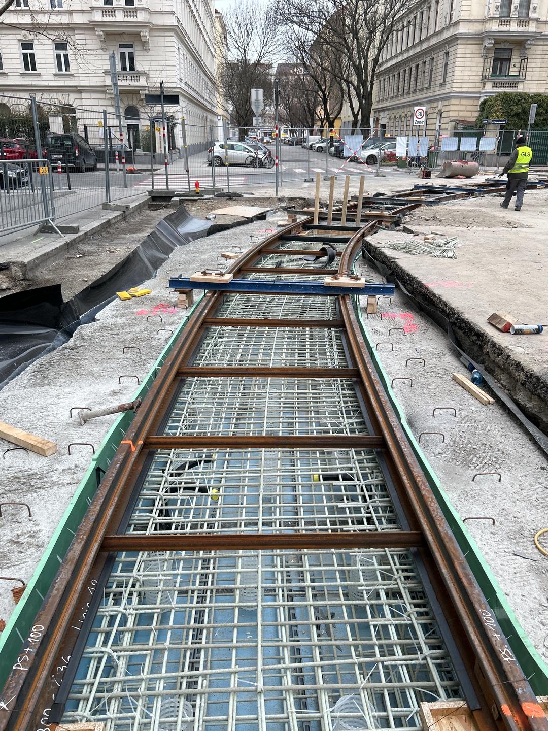 Baustelle mit sichtbaren Bewehrungsstäben aus Glasfaserkunststoff, darüber Gleise aus Stahl. Im Hintergrund sind Gebäude zu sehen.