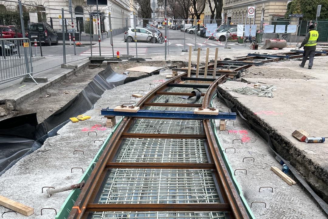 Baustelle mit sichtbaren Bewehrungsstäben aus Glasfaserkunststoff, darüber Gleise aus Stahl. Im Hintergrund sind Gebäude zu sehen.