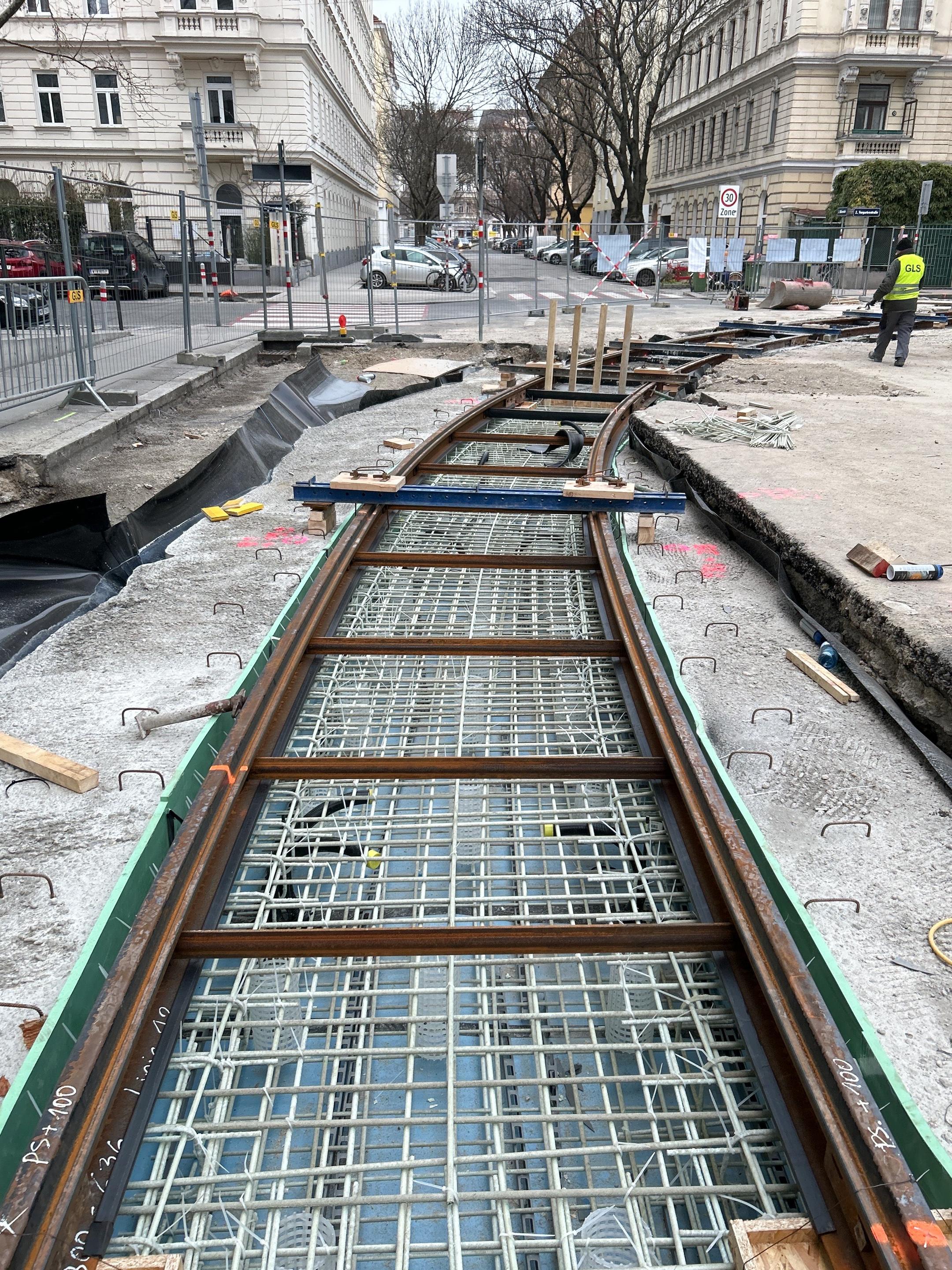Baustelle mit sichtbaren Bewehrungsstäben aus Glasfaserkunststoff, darüber Gleise aus Stahl. Im Hintergrund sind Gebäude zu sehen.