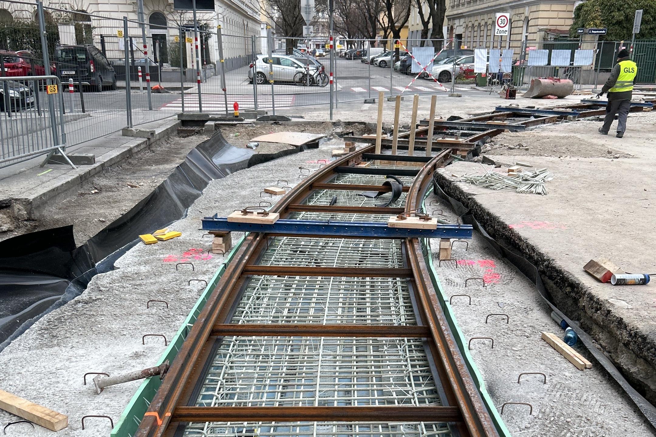 Baustelle mit sichtbaren Bewehrungsstäben aus Glasfaserkunststoff, darüber Gleise aus Stahl. Im Hintergrund sind Gebäude zu sehen.