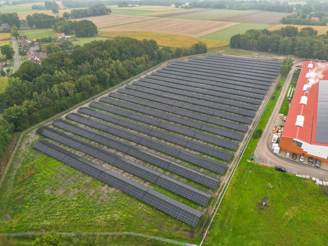 Luftaufnahme einer Photovoltaikanlage mit Feldern und Bäumen im Hintergrund, ein Industriegebäude schließt sich an.