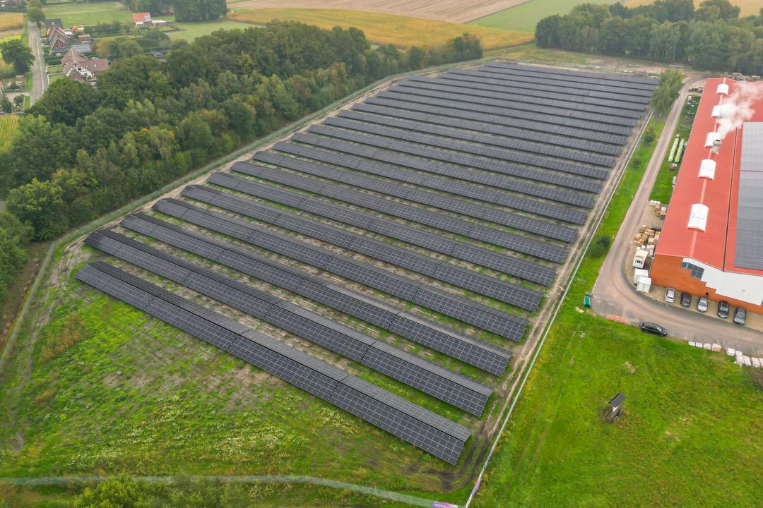 Luftaufnahme einer Photovoltaikanlage mit Feldern und Bäumen im Hintergrund, ein Industriegebäude schließt sich an.