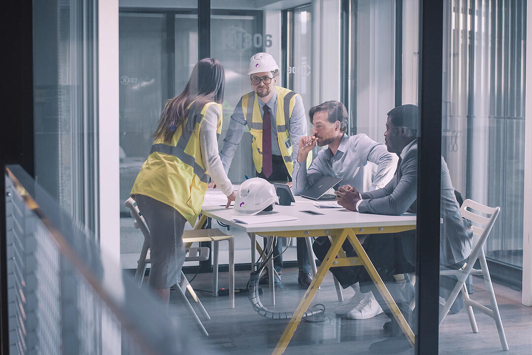 Projektteam mit Warnwesten und Bauhelmen bespricht Pläne und Unterlagen in einem modernen Besprechungsraum.